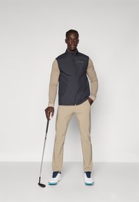 Menns ermeløse svarte vest over en beige langtermet skjorte, kombinert med beige bukser og hvit-blå golf sko, mens han holder en golfkølle.