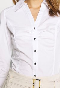 Vit skjorta med en figurnära design, som har en spetskrage, veck vid bysten och svarta knappar framtill.