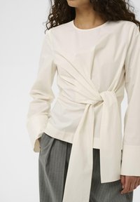 Valge pikakäistega särk, millel on sõlmega vöökoht, ümmargune kaelus ja kortsuvaba riie, sobitatud hallide triibuliste pükstega.