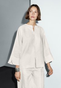 Blouse en lin gris clair avec un col rond, une fermeture à boutons sur le devant et des manches larges. Présente une texture subtile et une finition lisse.