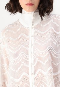 White lace blouse featuring a high collar, intricate wavy patterns, and button closures, with a sheer texture and decorative accents.