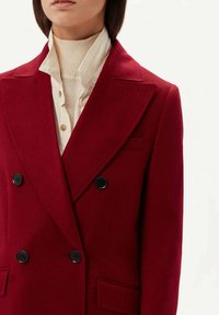 Röd ull dubbelknäppt kavaj med svarta knappar, spetsiga slag och två framfickor; lager på lager över en beige polo-tröja.