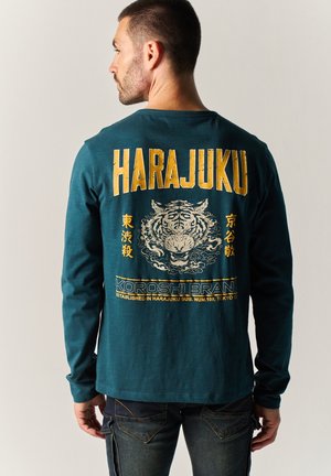 Hombre de espaldas vistiendo una camiseta de manga larga color verde azulado con texto amarillo "HARAJUKU" y un gráfico de tigre en la espalda.