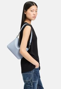 Jasnoniebieska skórzana torebka o luźnym fasonie, z srebrnymi elementami i pojedynczym paskiem, noszona na ramieniu z czarną bluzką i dżinsami.