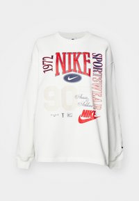 Witte sweatshirt met lange mouwen met rode en marineblauwe Nike-logo's, "1972," "SPORTSWEAR," "90," en decoratieve tekst op de voorkant.