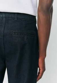 Dark denim trousers featuring a slim fit, a back pocket with a button closure, and a textured finish.