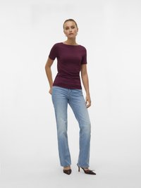 Žena stoji u kratkoj bordo majici s kratkim rukavima, svijetloplavim trapericama ravnog kroja i smeđim visokim štiklama sa špicom, na bijeloj pozadini.