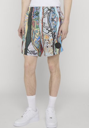 Mann trägt mehrfarbige, gemusterte Shorts mit abstrakten Designs, weiße Socken und weiße Turnschuhe, steht vor einem einfarbigen hellen Hintergrund.
