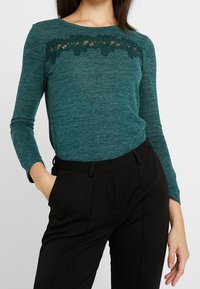 Turkusowa bluzka z długim rękawem z koronkowym detalem na torsie, w zestawieniu z czarnymi eleganckimi spodniami. Gładka tekstura i dopasowany krój.