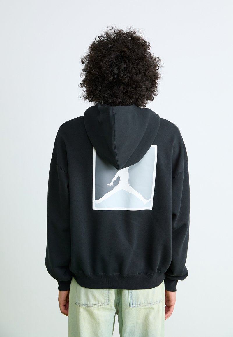 Black hoodie with a large white graphic of a jumping figure on the back. Features a drawstring hood and ribbed cuffs. Soft fabric texture.