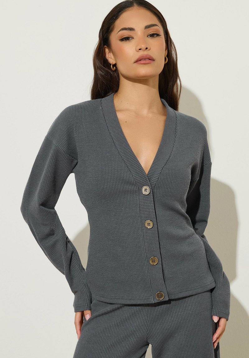 Femme portant un cardigan gris côtelé boutonné et un pantalon assorti, debout les mains dans les poches sur un fond clair.