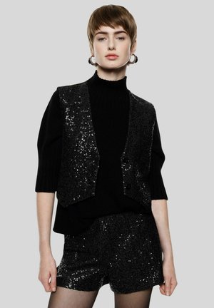 Donna con capelli corti che indossa un gilet e pantaloncini neri con paillettes sopra un dolcevita nero, in piedi davanti a uno sfondo grigio semplice.