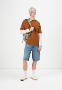 Brun t-shirt over en hvidstribet langærmet skjorte, denim shorts og beige sneakers. Grå crossover taske. Lys baggrund.