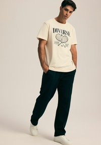 Kremowa koszulka z krótkim rękawem z grafiką tenisową oraz ciemnozielone szerokie spodnie, w połączeniu z białymi sneakersami. Prosty, casualowy design.