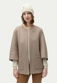 Beige ullblandning capa med rund hals, två framfickor och en subtil logotypdetalj. Bärs över en krämfärgad tröja och ljusbruna byxor.
