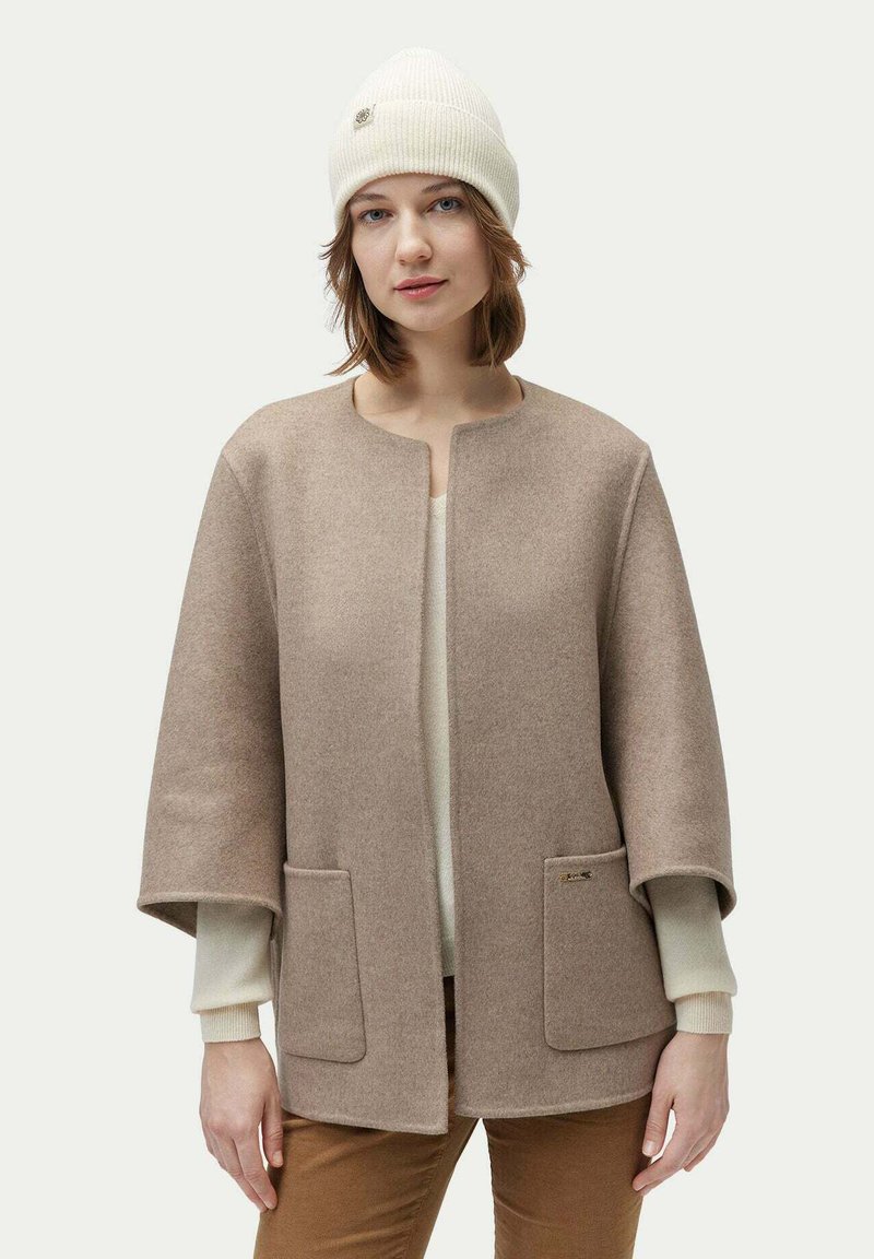 Beige ullblandning capa med rund hals, två framfickor och en subtil logotypdetalj. Bärs över en krämfärgad tröja och ljusbruna byxor.