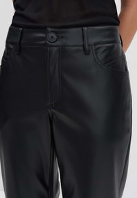 Close-up van zwarte neplederen broek met knoopsluiting, riemlussen en voorzakken, gedragen met een zwarte top.