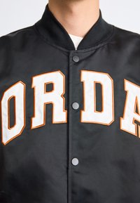 Person wearing a black varsity jacket with large white and orange outlined letters "ORDA" on the front and black snap buttons.