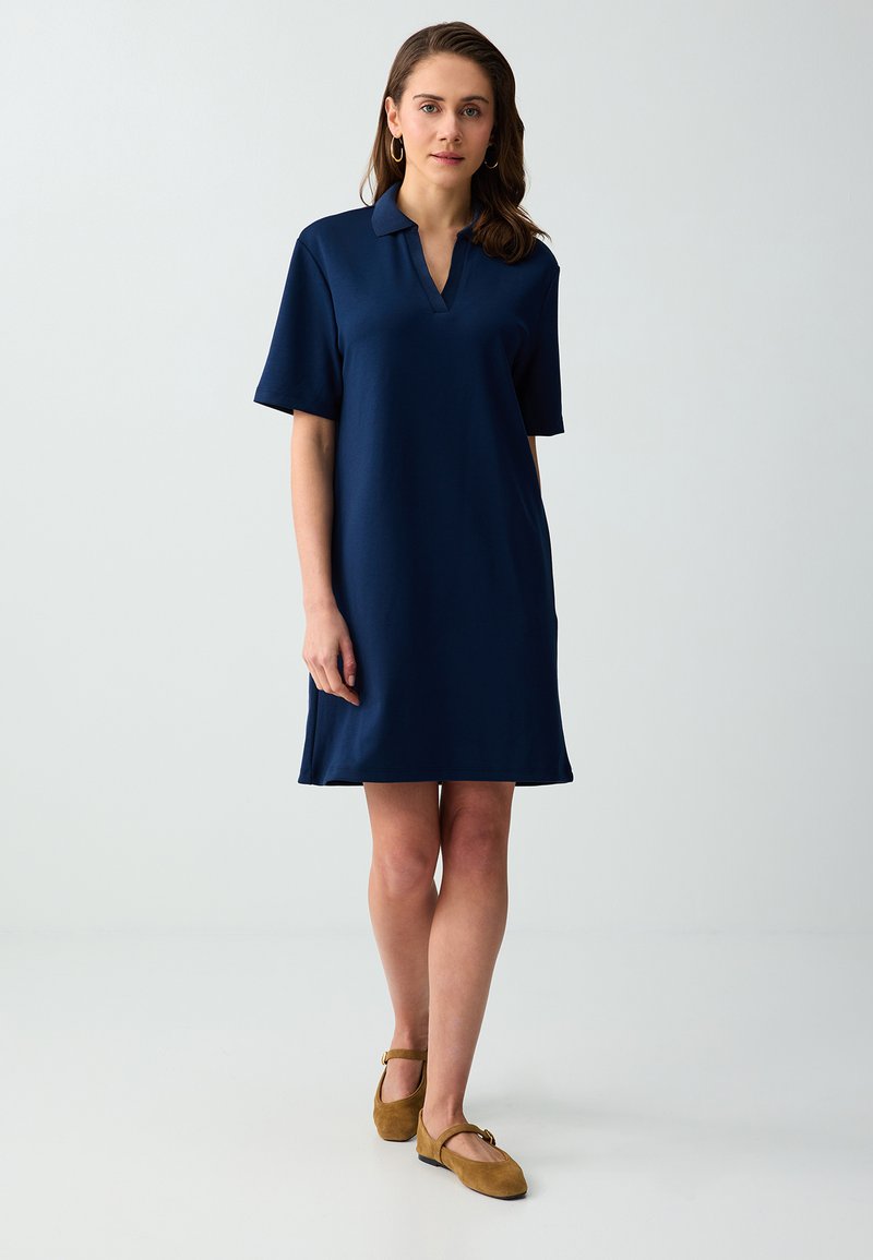 Femme portant une robe polo bleu marine à manches courtes et des ballerines marron à bride cheville, debout devant un fond clair uni.
