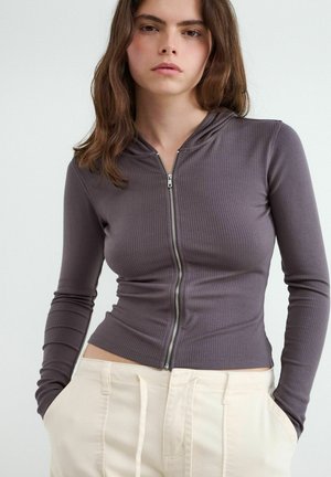 Mladá žena s hnedými vlasmi, oblečená v sivom rebrovanom mikinovom zipsu a krémových nohaviciach, ruky v vreckách, neutrálny background.