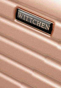Primer plano de equipaje rígido de color oro rosa con surcos horizontales y una etiqueta negra que muestra el nombre de la marca "WITTCHEN".