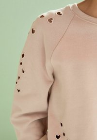 Felpa rosa chiaro con fori a forma di cuore sulle spalle, realizzata in tessuto morbido con scollatura girocollo classica e polsini a coste.