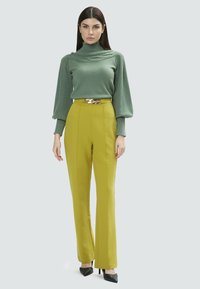 Maglione verde a collo alto lavorato a maglia con maniche a sbuffo, abbinato a pantaloni gialli a vita alta con cintura in catena dorata e tacchi neri.