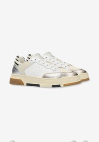 Zapatillas deportivas con parte superior de cuero blanco liso, detalles metálicos plateados y talón texturizado. Suela de goma gruesa con detalles en negro y marrón.
