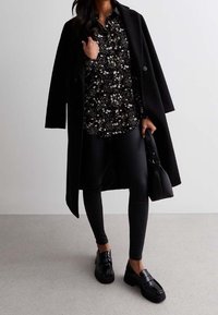 Musta villakangas takki, jonka päällä on kukkakuosiset musta-valkoiset paita, yhdistettynä mustiin leggings-housuihin ja kiiltäviin mustiin loafer-kengiin. Mukana on rakenteellinen käsilaukku.