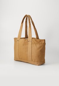 Tan canvas tote bag with dual handles, featuring side pockets and visible stitching details. Constructed from sturdy fabric with a soft texture.