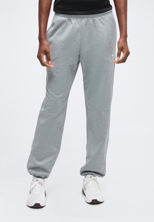 Personne portant un pantalon de jogging gris avec des chevilles élastiques et des chaussures de sport blanches, debout devant un fond clair uni.