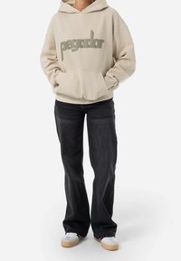 Felpa crema con la scritta "pegador" in lettere verdi, tasca frontale e cappuccio con coulisse regolabile. Abbinata a jeans neri a gamba larga e scarpe da ginnastica bianche.