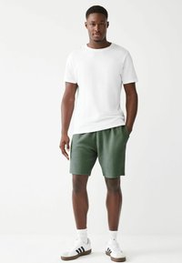 T-shirt bianca in cotone con scollo rotondo e maniche corte, abbinata a pantaloni corti verdi con texture e sneaker bianche con strisce nere.