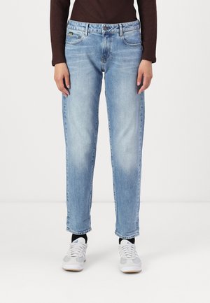 Personne portant un jean droit bleu clair, des chaussettes noires, des baskets blanches et un haut à manches longues marron foncé, debout devant un fond blanc.