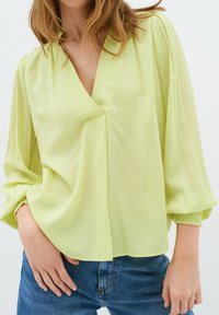 Blusa verde chiaro con scollo a V e maniche ampie e arricciate. Tessuto morbido con vestibilità rilassata, abbinata a jeans in denim blu.