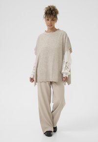 Beige strik poncho sweater med bølgemønster i kanten og subtile udsmykninger, lagd over en hvid top med blonder, parret med beige bukser.