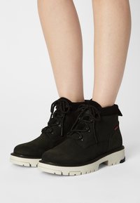 Black ankle boots made of textured suede with a white rubber sole, featuring black laces and metal eyelets. Slender shaft with a padded collar.