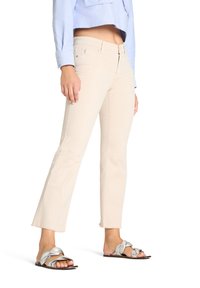 Mujer con jeans beige recortados, camisa azul clara recortada y sandalias planas metálicas plateadas, de pie frente a un fondo blanco.