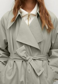 Lys grøn trenchcoat med en stor krave, front båndsløjfe, sidelommer og minimale syninger. Satyrlignende tekstur og oversized design.