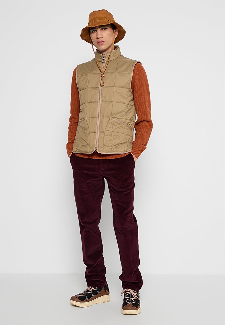 Gilet trapuntato beige con zip frontale e tasche, maglione ruggine sotto, pantaloni in corduroy bordeaux, sneakers marroni e nere, cappello a secchiello beige.