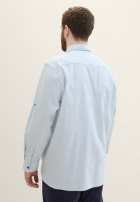 Mann mit kurzen braunen Haaren und Bart, der ein hellblaues langärmliges Hemd mit Knöpfen trägt, von hinten vor einem schlichten Hintergrund betrachtet.