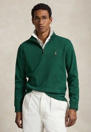 Homme portant des lunettes rondes, un pull vert à fermeture éclair jusqu'au quart sur une chemise blanche à col, et un pantalon blanc plissé sur fond neutre.