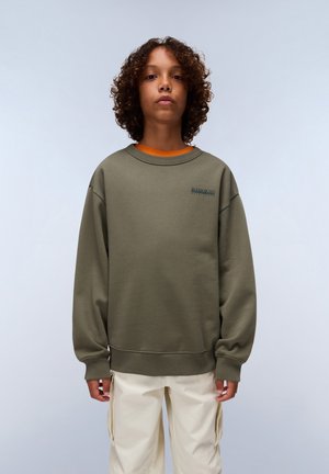 Sudadera verde oliva con cuello redondo y puños de canalé, que presenta un pequeño logotipo impreso en el pecho, llevada sobre una camisa naranja.