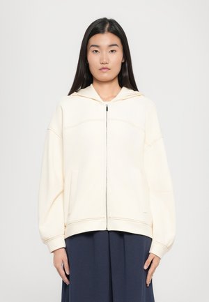Light cream hooded sweatshirt with a front zipper, two side pockets, and loose sleeves, featuring visible stitching along the seams.