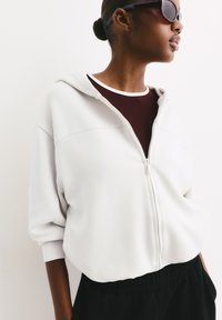 Biely mikina na zips s kapucňou, vyrobená z mäkkého materiálu, so zúženými rukávmi a voľným strihom, preložená cez burgundské tričko a čierne nohavice.