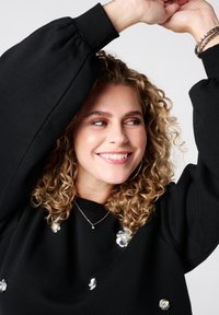 Zwarte sweatshirt met pofmouwen, versierd met doorzichtige bloemapplicaties. Model glimlacht met opgetilde armen, waarmee de ontwerpelementen worden getoond.