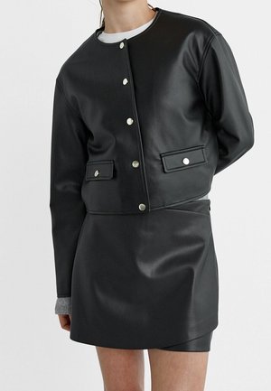 Femme portant une veste noire en cuir synthétique boutonnée et une jupe assortie avec des manches grises visibles aux poignets, sur un fond uni.