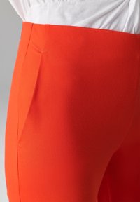 Rode rekbare broek met een gladde textuur, een getailleerde pasvorm en subtiele plooidetails aan de zijkant. De stof is lichtgewicht en elastisch.