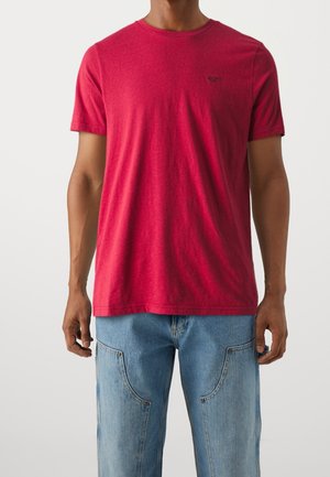 Rotes Baumwoll-T-Shirt mit Rundhalsausschnitt, kurzen Ärmeln und einem dezenten Logo-Akzent auf der Brust, kombiniert mit hellblauen weiten Jeans.