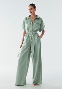 Svetlo zelena, kratka kombinacija s kratkimi rokavi z zapiranjem na gumbe, žepoma na prsih in pasom na zavezovanje. Obsežen kroj z mehko teksturo.